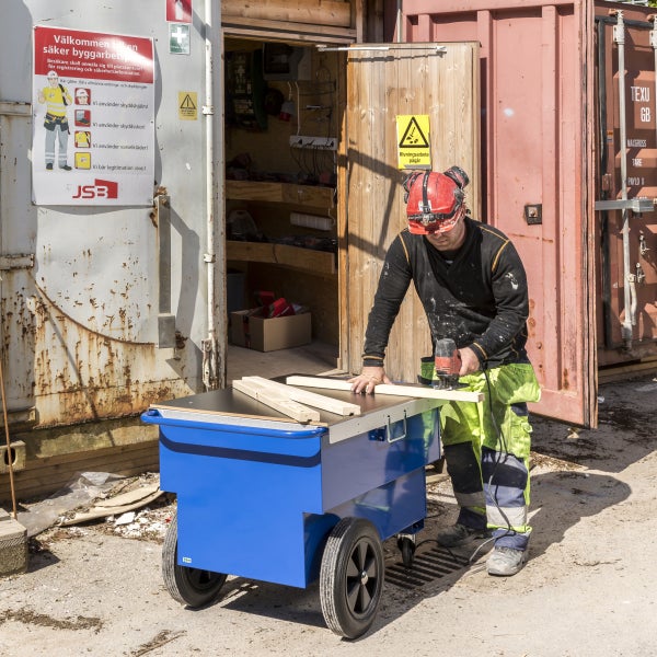 Ein Arbeiter schneidet Holz auf einem blauen Werkzeugwagen vor einem Baucontainer.