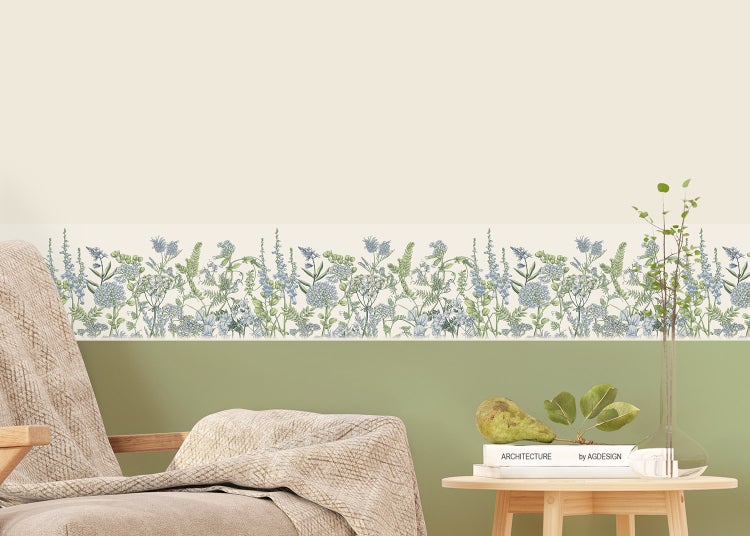 Tapetenbordüre mit floralem Wiesenblumenmuster an einer zweifarbigen Wand in einem hellen Raum mit Sessel und hölzernem Beistelltisch.