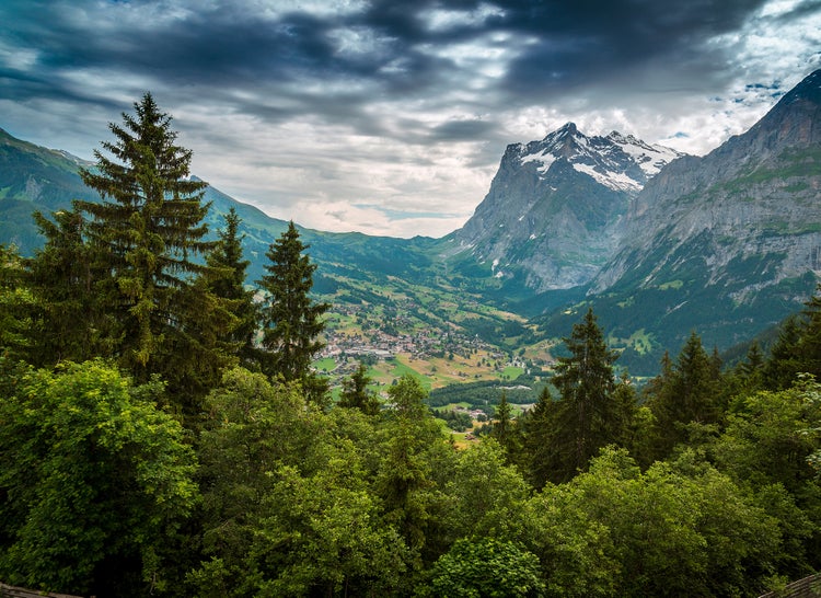 Wandbild Gebirgslandschaft mit verschneiten Gipfeln, Tal mit Dorf und Nadelwald unter bewölktem Himmel.