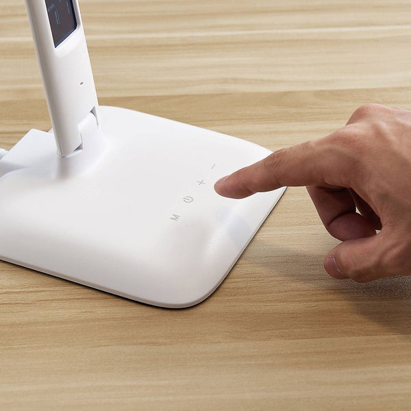 Hand bedient das berührungsempfindliche Bedienfeld am Sockel einer weißen Schreibtischlampe auf einem Holztisch.