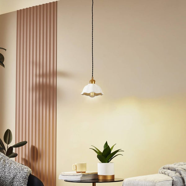 Pendelleuchte mit weißem gewelltem Schirm, goldener Fassung und schwarzem Kabel über einem Beistelltisch mit Pflanze in modernem Wohnzimmer.