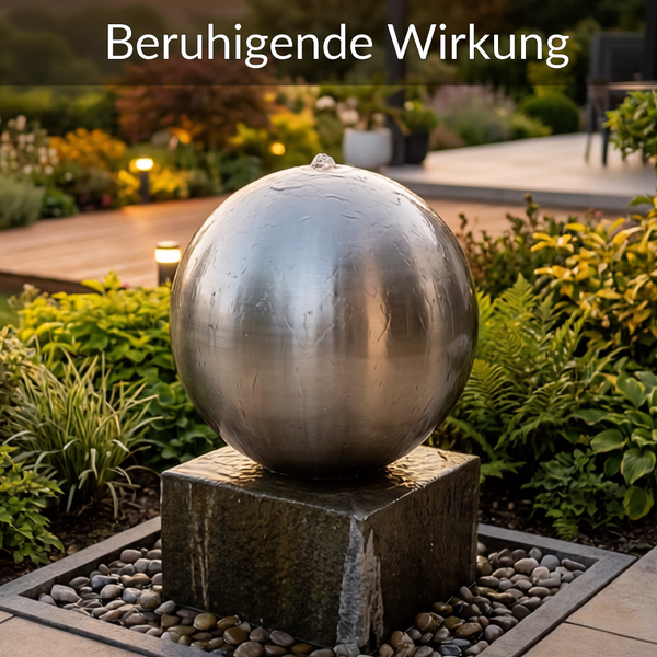 Kugelbrunnen aus Edelstahl auf einem dunklen Steinsockel in einem Garten mit Pflanzen und Kieselsteinen.
