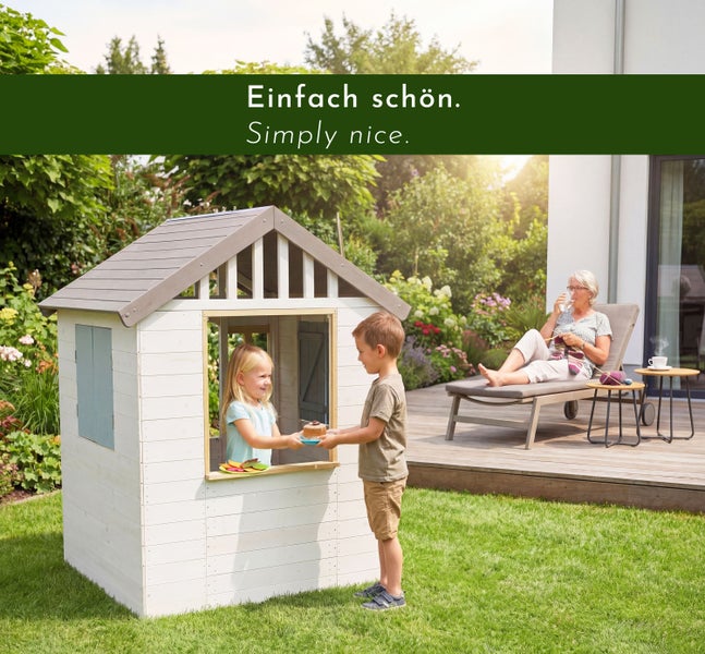 Zwei Kinder spielen mit einem weißen Garten-Spielhaus aus Holz auf einer Rasenfläche neben einer Holzterrasse in einem hellen Garten.