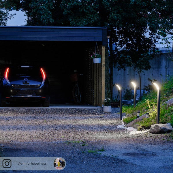 Drei moderne LED-Wegleuchten beleuchten eine Kiesauffahrt vor einem Carport mit Auto in der Dämmerung.
