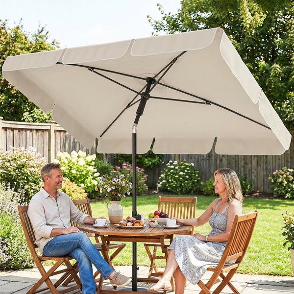 Ein Paar sitzt an einem Holztisch unter einem großen, rechteckigen Gartensonnenschirm mit Volant in einem hellen Garten.