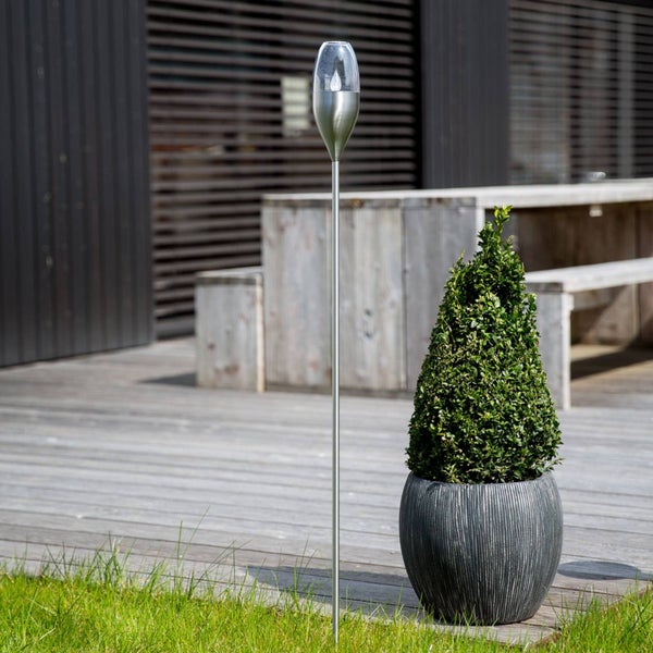 Hohe Solarfackel aus Edelstahl im Garten neben einem Pflanzkübel auf einer Holzterrasse.
