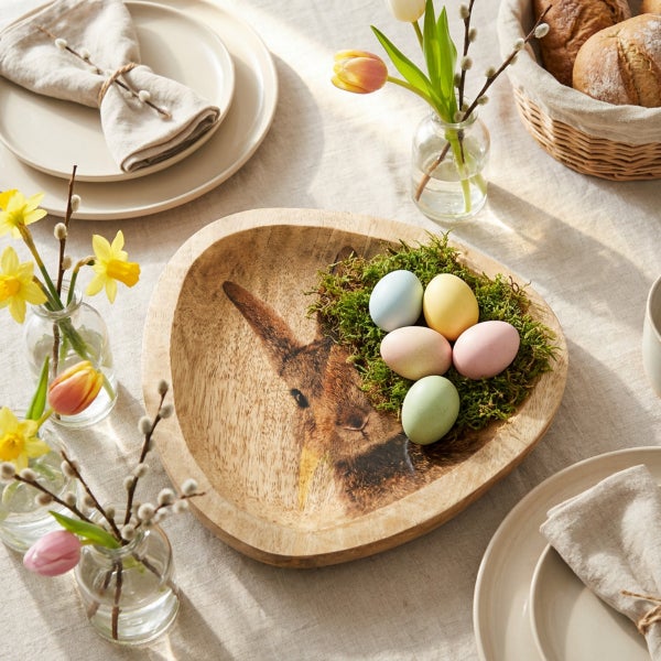 Holzschale in Hasenform mit Osterdekoration, Moos und bunten Ostereiern auf einem gedeckten Tisch.