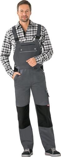Mann in grauer Latzhose mit schwarzen Kniebesätzen und einem schwarz-weiß karierten Hemd.