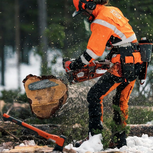 Eine Person mit Schutzkleidung sägt mit einer Kettensäge einen Baumstamm. Eine Axt liegt im Schnee.