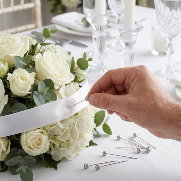 Hand steckt Stecknadel in ein weißes Satinband, das um einen Blumenstrauß aus Rosen und Hortensien für eine festliche Tischdekoration gelegt ist.