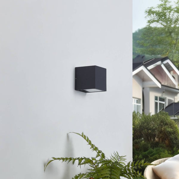 Dunkle quadratische Außenwandleuchte an einer Fassade, im Hintergrund Garten und Wohnhaus.