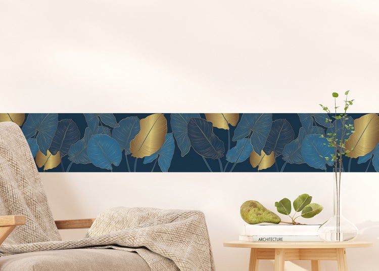 Tapetenbordüre mit tropischem Blattmuster in Blau und Gold an einer Wand über einem Sessel und einem Beistelltisch.