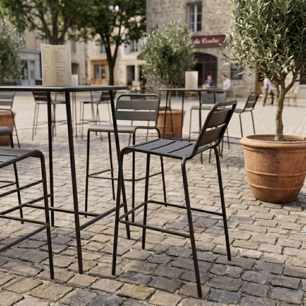 Gartenbarstühle und Stehtische aus Metall auf einer gepflasterten Terrasse mit Olivenbäumen in Tontöpfen.