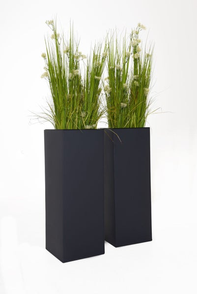 Zwei hohe rechteckige schwarze Pflanzgefäße mit Kunstgras und weißen Blüten.
