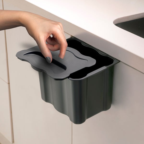 Eine Hand öffnet den Deckel eines kleinen schwarzen Bio-Abfalleimers aus Kunststoff, der an einem Küchenschrank befestigt ist.