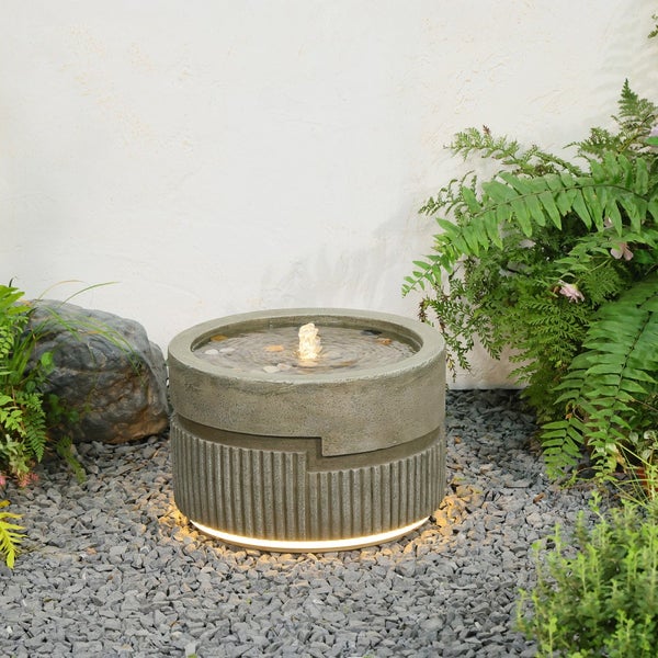Runder Gartenbrunnen in Steinoptik mit geriffeltem Sockel und LED-Beleuchtung am Boden, auf Kies stehend zwischen Gartenpflanzen.