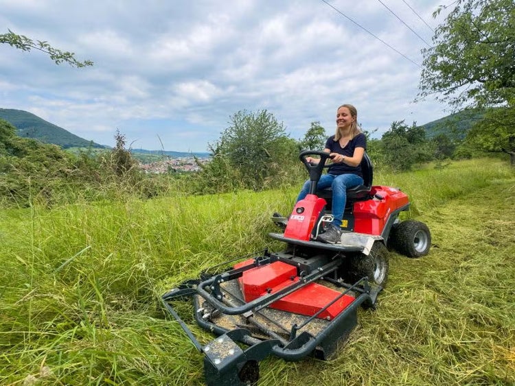 Eine Frau bedient einen ERCO Hochgrasmäher auf einer Wiese mit hohem Gras in einer hügeligen Landschaft.
