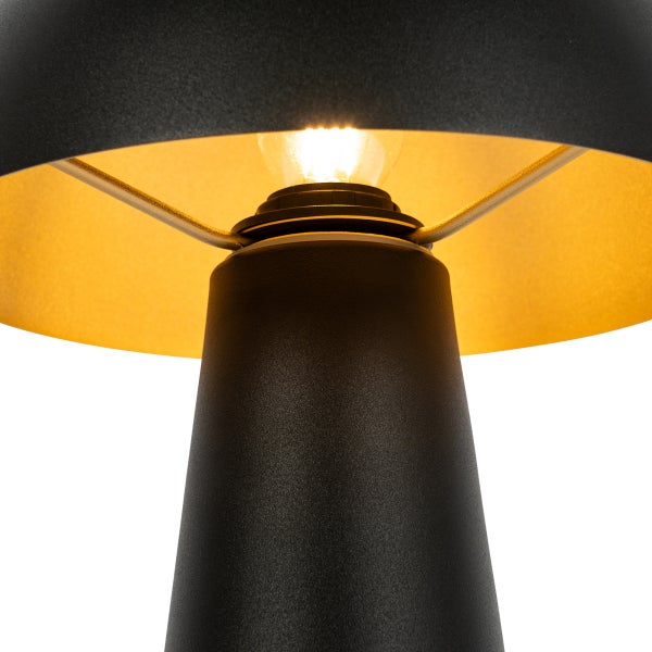 Detailaufnahme einer schwarzen Tischlampe aus Metall mit goldfarbener Innenseite und leuchtendem Leuchtmittel.