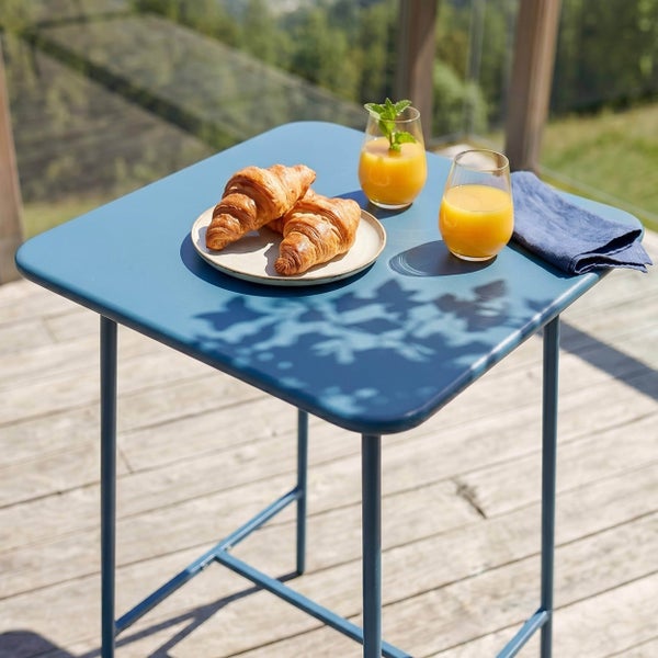 Blauer quadratischer Bistrotisch aus Metall auf einer hellen Holzterrasse mit Croissants, Gläsern Saft und einer Stoffserviette.