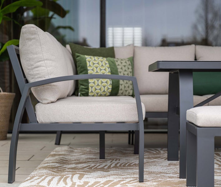 Modernes Outdoor-Loungeset mit dunkelgrauem Metallgestell, hellen Polstern und grünen Dekokissen auf einem gemusterten Teppich.