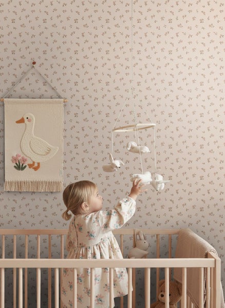 Vliestapete mit floralem Muster in einem hellen Kinderzimmer mit Gitterbett aus Holz, Wandteppich mit Gans-Motiv und Mobile.