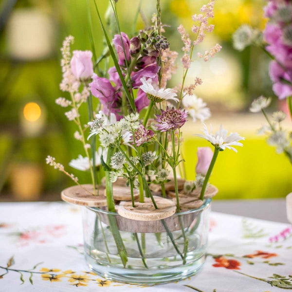 Blumenarrangement in einer niedrigen Glasvase mit einem Blumenhalter aus Holzscheiben auf einem Tisch.