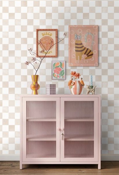 Hellbeig-weiß karierte Tapete in einem Wohnraum hinter einer rosa Metallvitrine mit geriffelten Glastüren und verschiedenen Wandbildern.