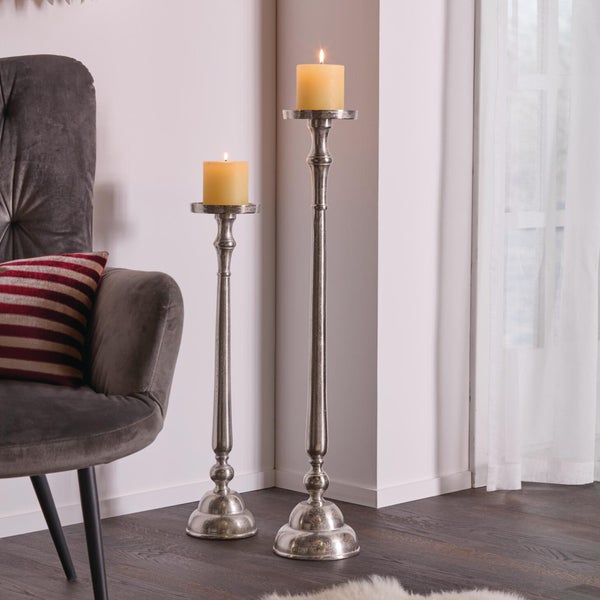 Zwei metallene Kerzenständer in unterschiedlicher Höhe mit brennenden Stumpenkerzen neben einem Sessel in einem Wohnraum.