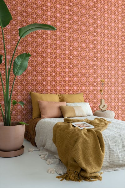 Schlafzimmer mit floral gemusterter Tapete im Retro-Stil, einem Bett mit verschiedenen Kissen und einer großen Zimmerpflanze im Topf.