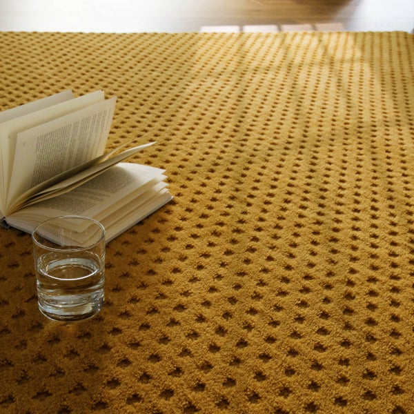 Senfgelber strukturierter Teppich mit Wabenmuster, auf dem ein aufgeschlagenes Buch und ein Glas Wasser liegen.