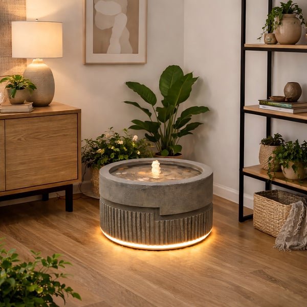 Runder Zimmerbrunnen in Beton-Optik mit LED-Beleuchtung am Sockel und im Wasser in einem gemütlichen Wohnzimmer mit Holzdekor und Zimmerpflanzen.
