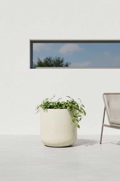 Moderner runder Pflanzkübel in Steinoptik mit grüner Pflanze auf einer hellen Terrasse vor einer weißen Wand mit Fenster und Gartenstuhl.