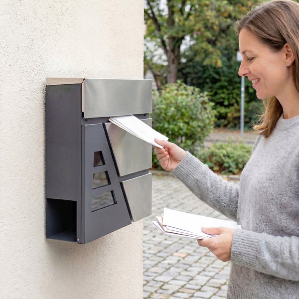 Eine Frau wirft Post in einen anthrazitfarbenen Wandbriefkasten mit Edelstahl-Elementen an einer hellen Hauswand.