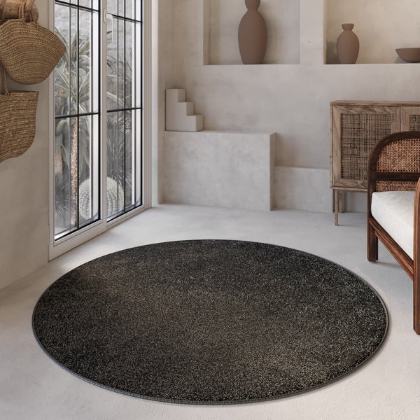 Runder, dunkel gesprenkelter Teppich in einem hellen, minimalistischen Raum mit Holzmöbeln und neutraler Dekoration.