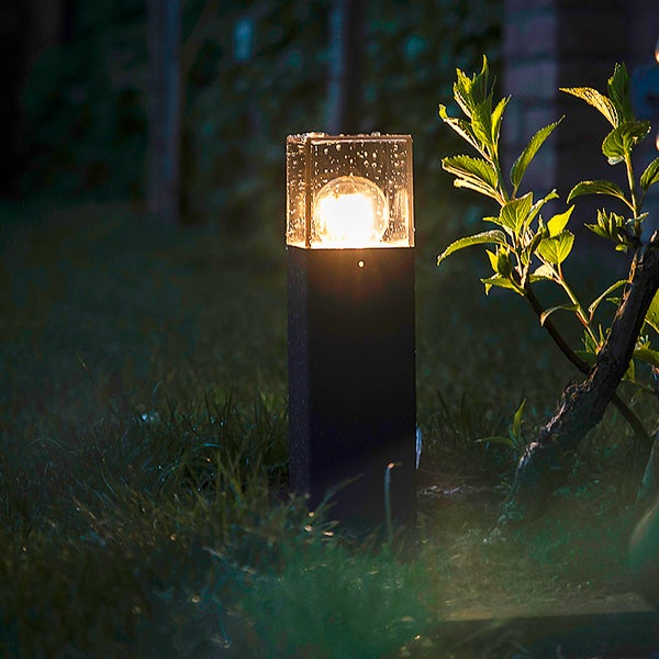 Moderne Außen-Sockelleuchte im Garten bei Nacht, warmes Licht, transparenter Schirm mit Wassertropfen, schwarzes Gehäuse.