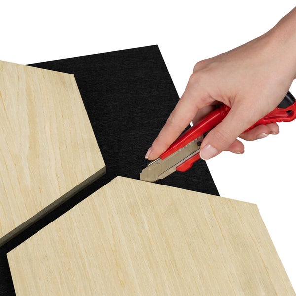Eine Hand schneidet mit einem roten Cuttermesser das schwarze Trägermaterial eines hexagonalen Akustikpaneels in Holzoptik zu.