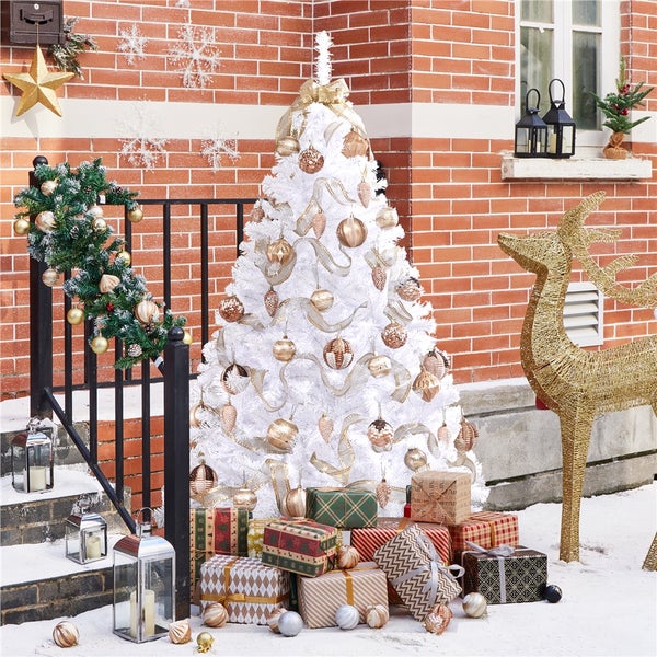 Dekorierter, weißer Weihnachtsbaum mit goldenem Schmuck neben Geschenken, Laternen und einem goldenen Rentier vor einem Backsteingebäude.