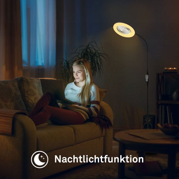 Frau sitzt auf einem Sofa mit Laptop, beleuchtet von einer Stehleuchte mit Nachtlichtfunktion in einem dunklen Wohnzimmer.