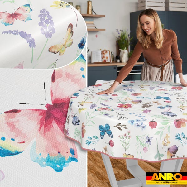 Eine Frau breitet eine runde Wachstuch-Tischdecke mit buntem Schmetterlings- und Blumenmuster in einer Küche aus. ANRO Logo.