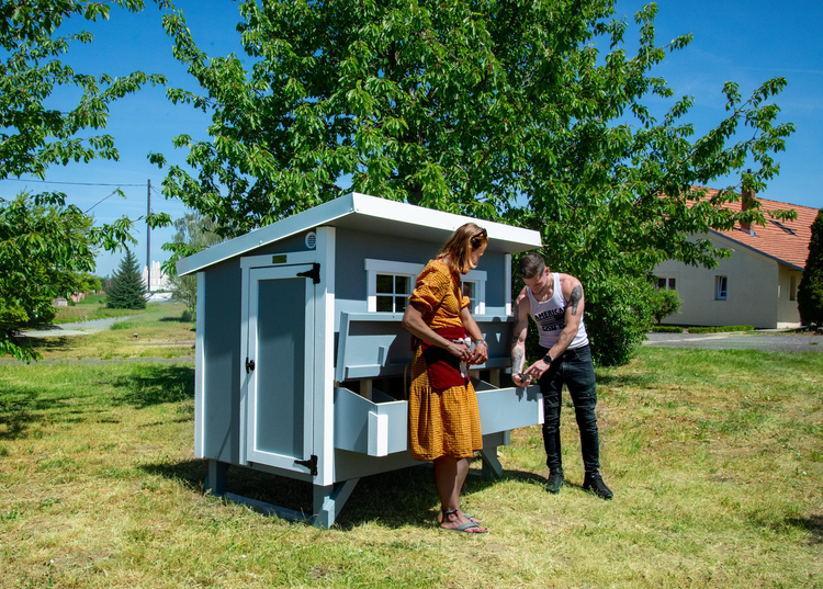 Moderner Hühnerstall aus Holz in Grau und Weiß mit Pultdach, Fenstern und ausziehbaren Nistkästen in einem Garten mit zwei Personen.
