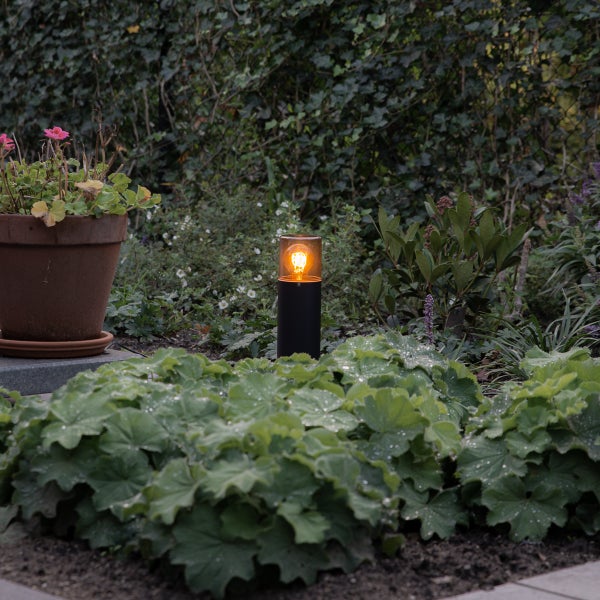 Schwarze, zylindrische Sockelleuchte mit warmem Licht in einem bepflanzten Gartenbeet neben einem Blumentopf aus Ton.