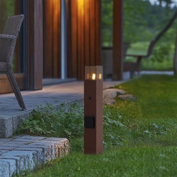 Moderne Wegeleuchte in Rostoptik mit integriertem Leuchtmittel und Steckdose auf einer Rasenfläche neben einer Steinterrasse.