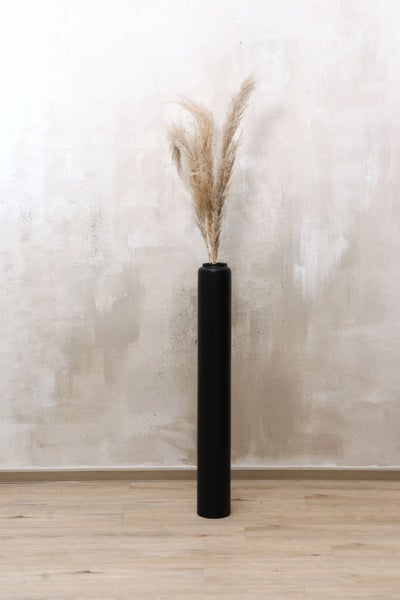 Hohe schwarze Bodenvase in Zylinderform mit getrocknetem Pampasgras auf Holzboden vor einer strukturierten Wand.