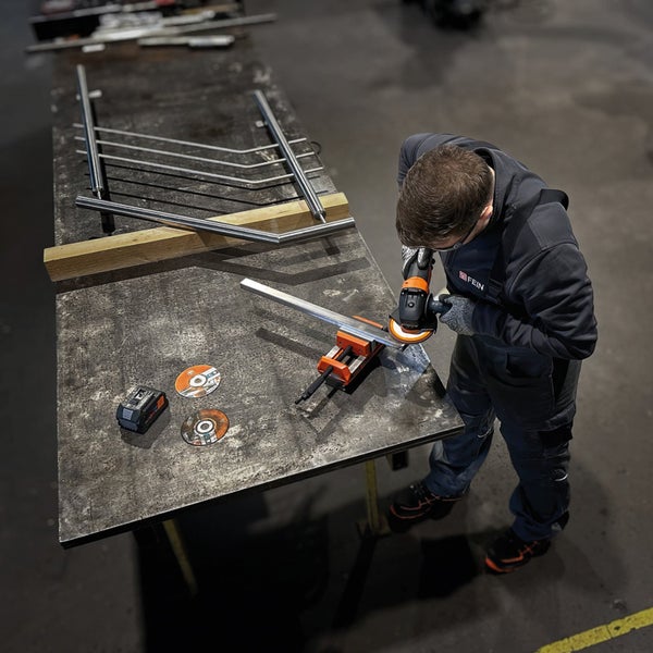 Handwerker mit FEIN Logo bearbeitet ein Metallprofil mit einem FEIN Akku-Winkelschleifer an einem Schraubstock auf einer Werkbank in einer Werkstatt.
