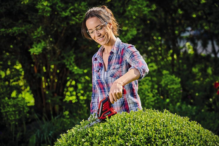 Eine Frau mit Schutzbrille stutzt mit einer Einhell Akku-Gras- und Strauchschere einen runden Busch im Garten.