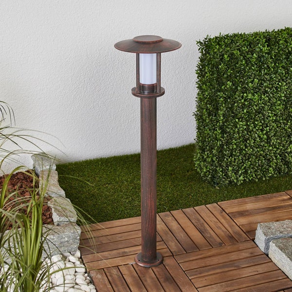 Wegeleuchte in Kupferoptik mit zylindrischem Diffusor auf einer Holzterrasse in einem Garten mit Hecke und Rasenfläche.