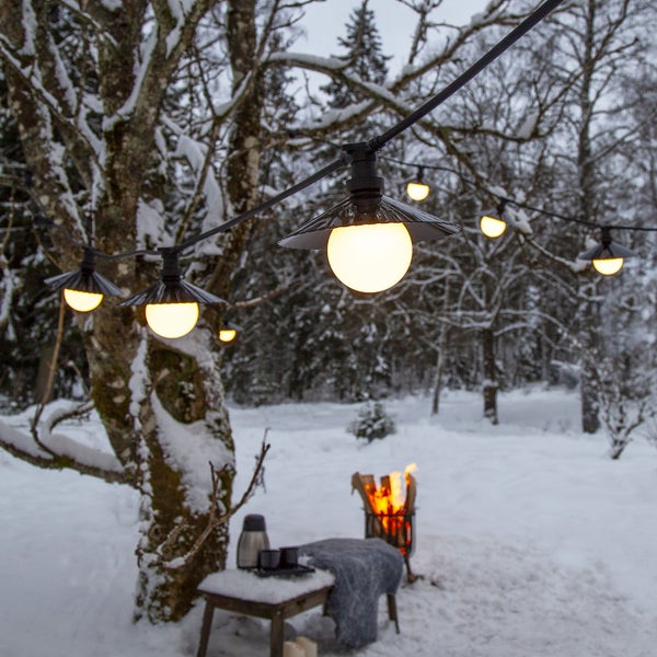 Lichterkette mit Lampen im Schnee, Tisch mit Getränken und Feuerkorb im Hintergrund