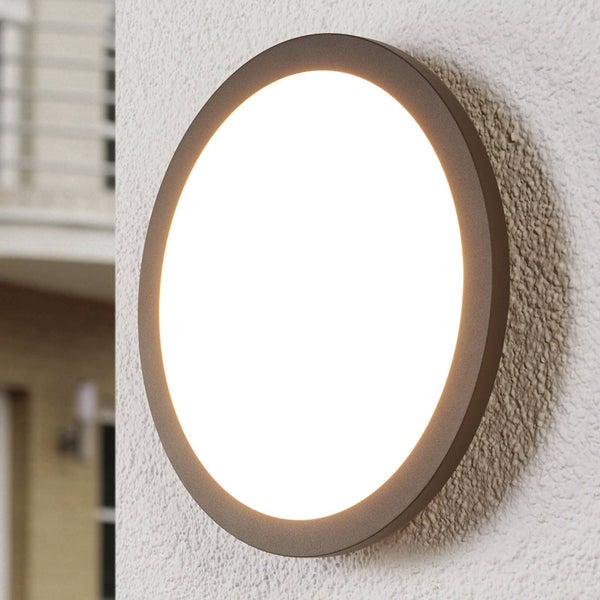 Runde LED-Außenwandleuchte mit dunklem Metallrahmen, montiert und eingeschaltet an einer strukturierten Wand.