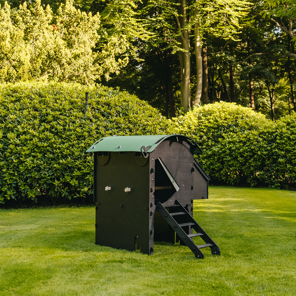 Moderner Hühnerstall aus Kunststoff mit grünem Bogendach und Leiter auf einer Rasenfläche im Garten umgeben von Bäumen und Sträuchern.