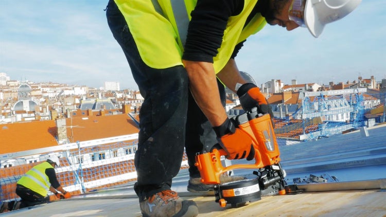 Handwerker bei der Arbeit auf einem Dach mit einem Druckluftnagler, im Hintergrund ein Stadtpanorama mit Ziegeldächern.